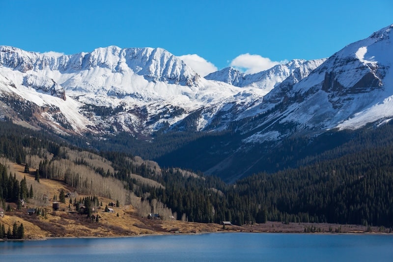 Scenic view of snow-covered Colorado mountains with pine trees and a lake in the foreground.
