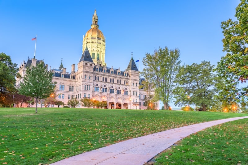 The Connecticut State Capitol in Hartford at dusk, with a golden dome and American and state flags, surrounded by a lush green lawn and autumn trees.