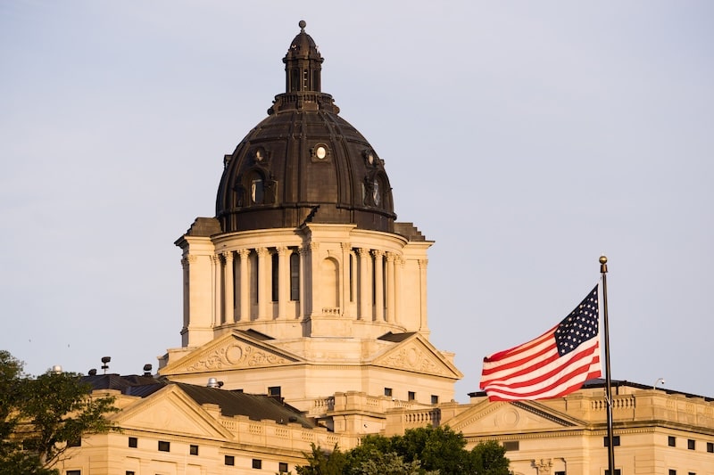 Sunrise view of the South Dakota State Capitol with an American flag waving in the foreground.
