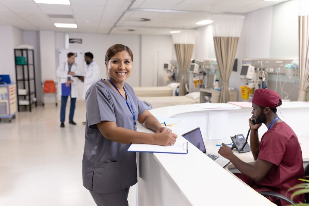 Healthcare professional in scrubs writing on a clipboard at a hospital nurses’ station.
